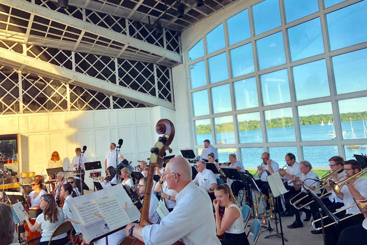 Inside the Lake Harriet band shell with Kenwood Orchestra performing on stage and the bright blue water sparkling through the windows with several sail boats floating.